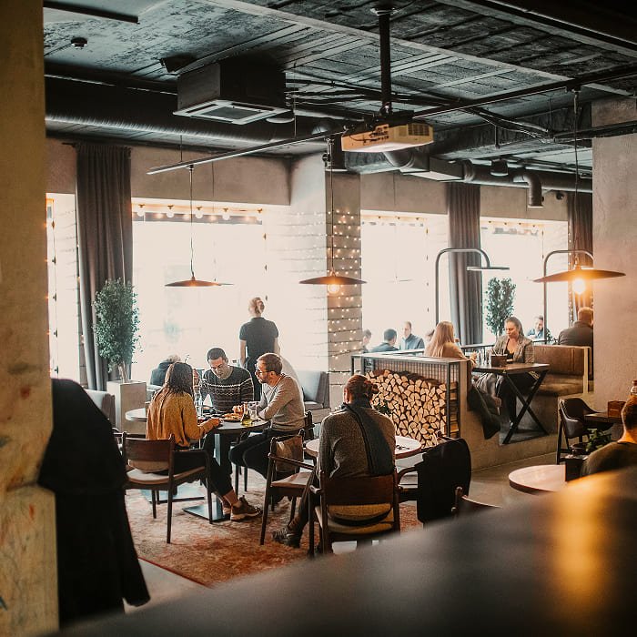 Svečiai sėdi prie stalų šviesioje restorano salėje prie didelių langų